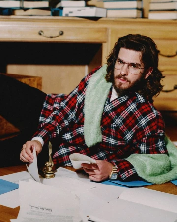 Andrew Garfield wearing glasses and a red plaid jacket reclining on a wooden floor surrounded by paperwork in front of a desk.