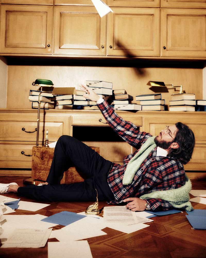 Andrew Garfield wearing a plaid blazer and eyeglasses lies on a wooden floor reaching toward a flying paper airplane.