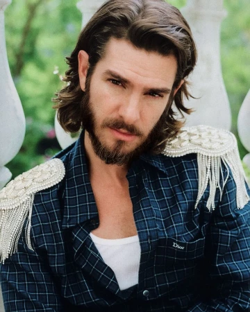 Portrait of Andrew Garfield wearing a navy plaid shirt with beaded fringe epaulettes outdoors.