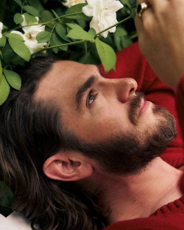 Close-up profile of Andrew Garfield lying among white roses and green foliage wearing a red sweater.