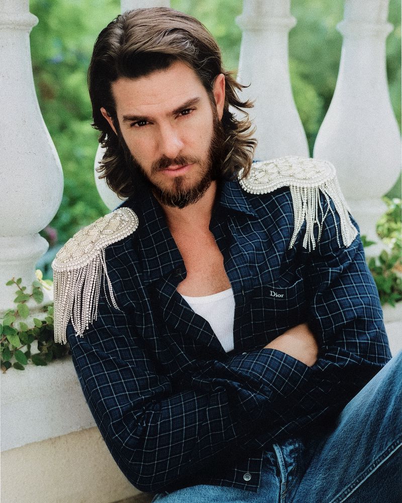 Actor Andrew Garfield sitting with arms crossed wearing a dark plaid shirt with ornate crystal fringe epaulettes by a white balustrade.