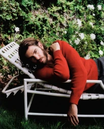 Andrew Garfield reclining on a white metal lawn chair wearing a red sweater in a garden setting.