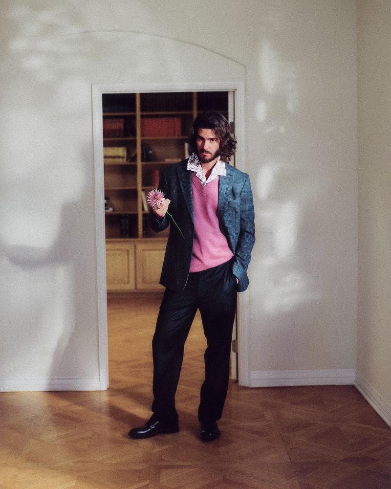 Andrew Garfield posing in a doorway wearing a charcoal blazer, pink sweater, and dark trousers while holding a pink flower.