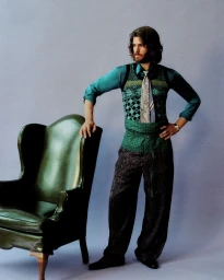 Andrew Garfield posing in a green patterned vest and patterned trousers next to a leather armchair against a light purple background