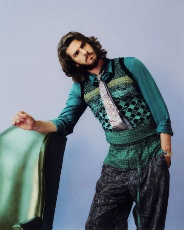 Andrew Garfield leaning against a green armchair wearing a patterned knit vest, teal shirt, and paisley trousers in a studio.