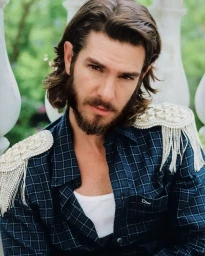 Actor Andrew Garfield with long hair and a beard wearing a dark plaid shirt with embellished shoulder appliques