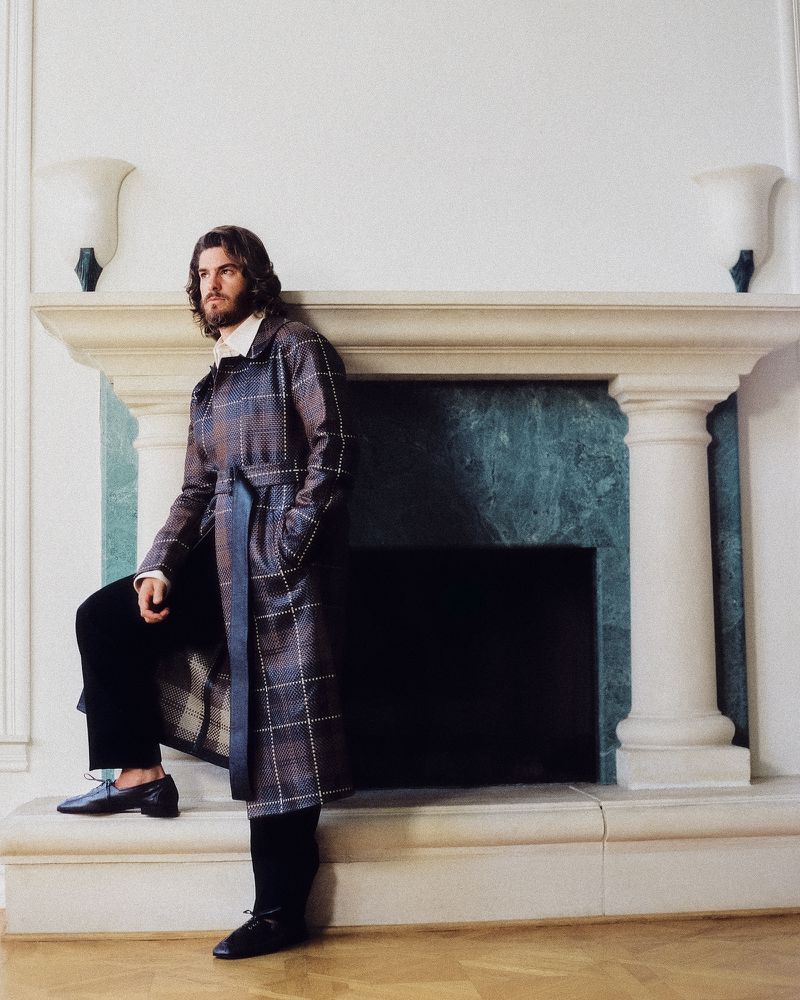 Actor Andrew Garfield wearing a belted plaid coat, standing beside a grand stone fireplace mantle.