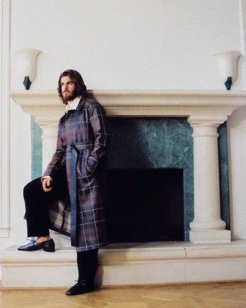 Actor Andrew Garfield wearing a belted plaid coat, standing beside a grand stone fireplace mantle.