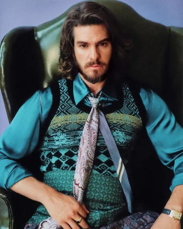Andrew Garfield seated in a dark green leather armchair wearing a teal shirt, knit sweater vest, and paisley tie.