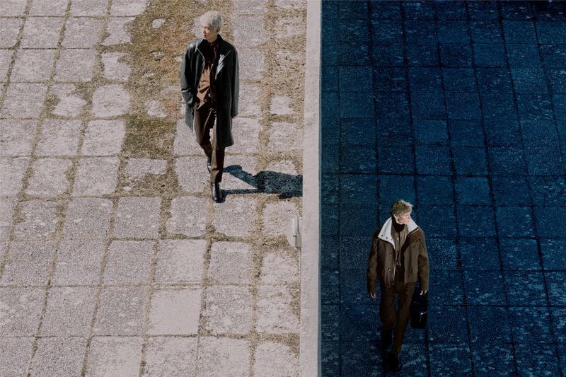 High angle shot of two male models in designer outerwear walking on split stone paved surfaces in contrasting light and shadow.