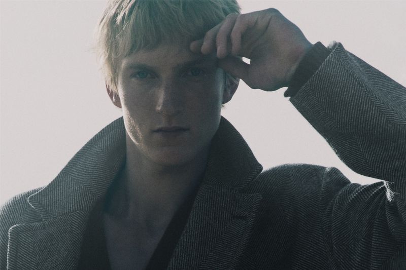 Pierrick Grégoire wearing a textured grey wool coat with hand raised to forehead in a fashion portrait.