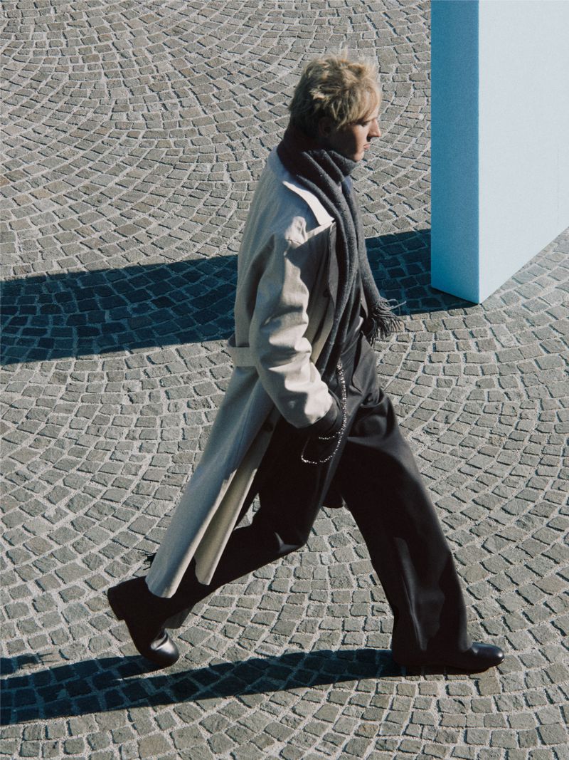 High-angle view of a man walking in a beige trench coat and wide-leg trousers across a cobblestone pavement.