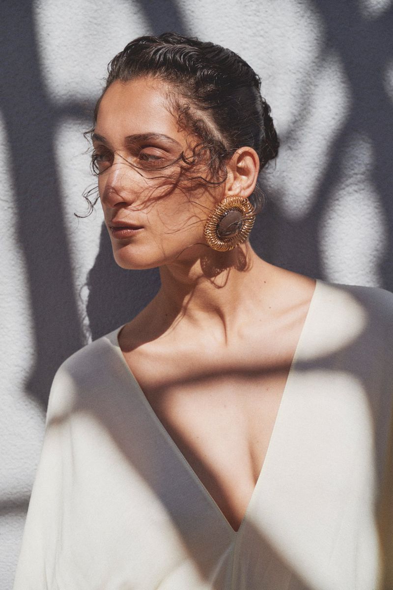 Portrait of a stylish adult female with elaborate earrings and a V-neck top, posing in front of a wall with geometric shadows.