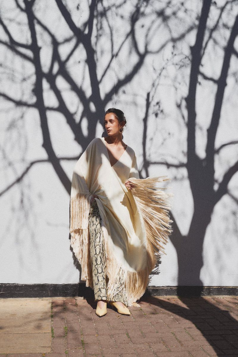 Adult woman poses in flowing white shawl with fringe and patterned pants. Tree shadows form pattern on white wall backdrop.