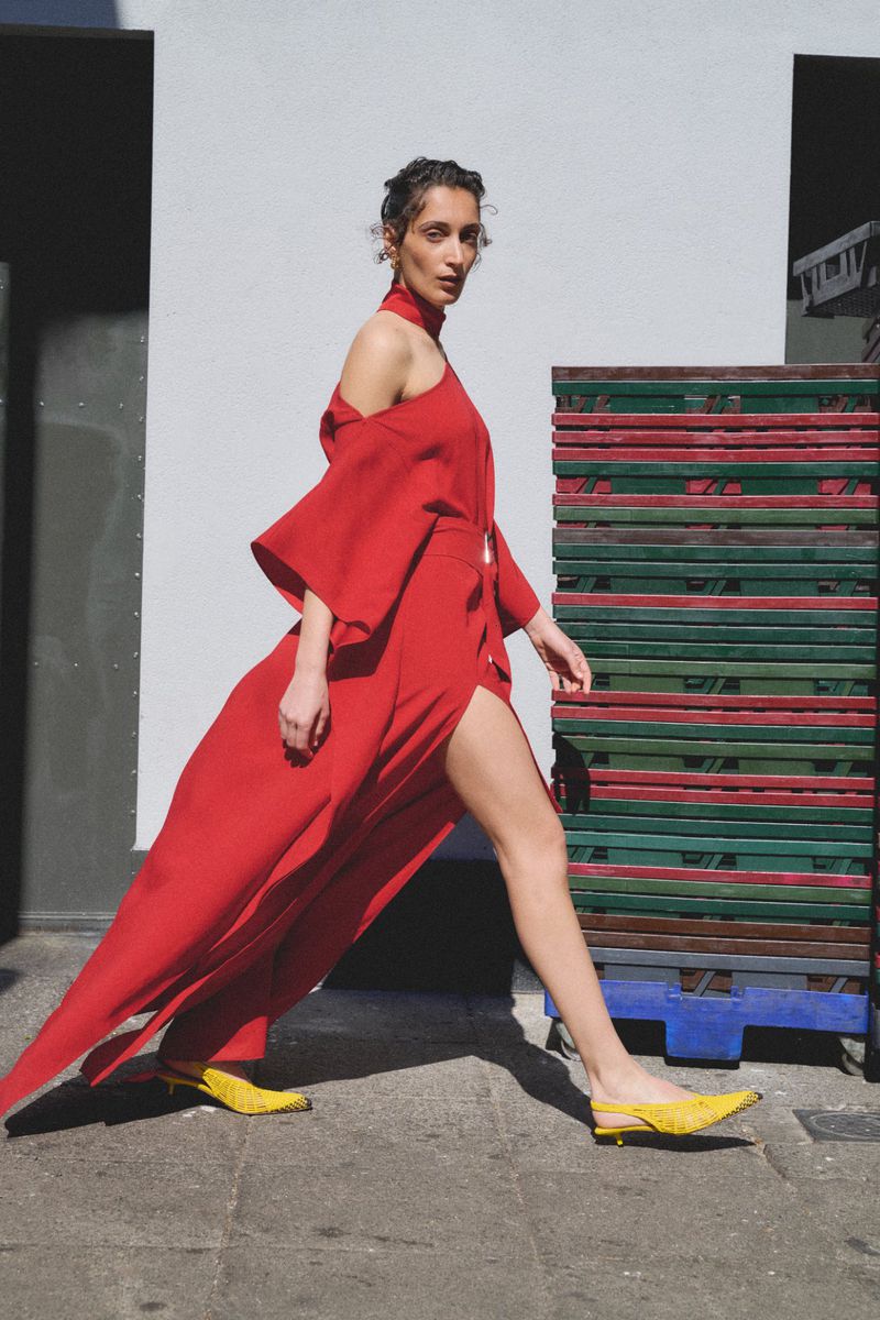 Woman in a red dress walking outdoors, showcasing high fashion against a neutral backdrop with vibrant yellow shoes and a stack of pallets.