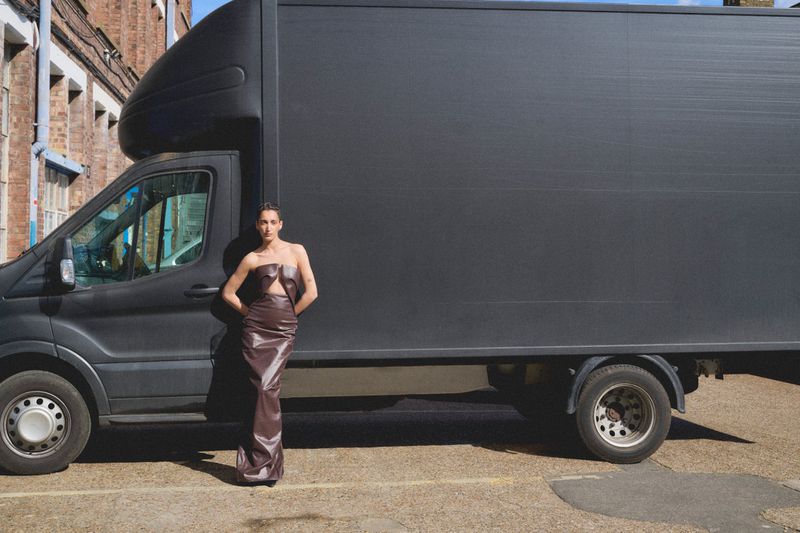 Adult female fashion model poses next to a large black delivery van in a brown strapless dress, creating a striking contrast in an urban setting.