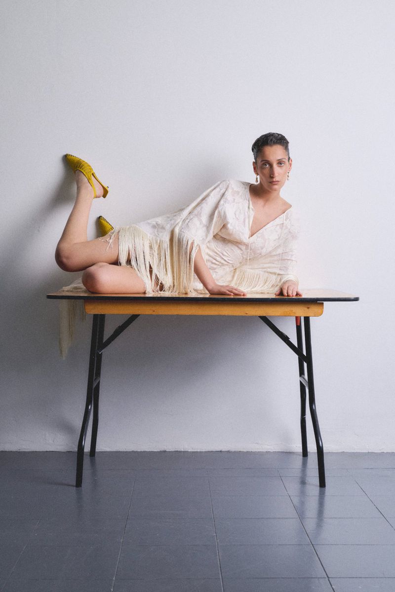 A woman with a short haircut lies on a folding table wearing a fringed white dress and yellow shoes in a minimalist studio setting.