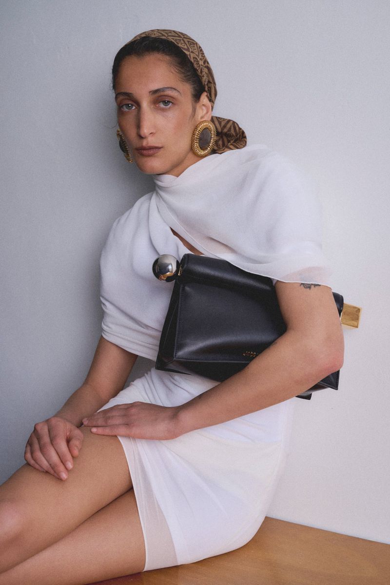 Stylish woman in white dress poses with a black handbag and patterned headscarf in a studio, showcasing fashion and elegance.
