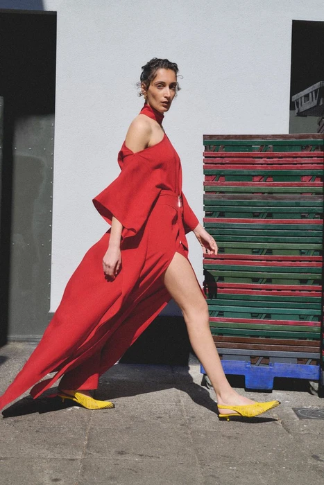 Model wearing a vibrant red dress and yellow shoes walking past stacked crates on an urban street.