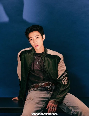 Jay Park seated wearing a green and beige Diesel jacket and distressed denim jeans against a deep blue backdrop.