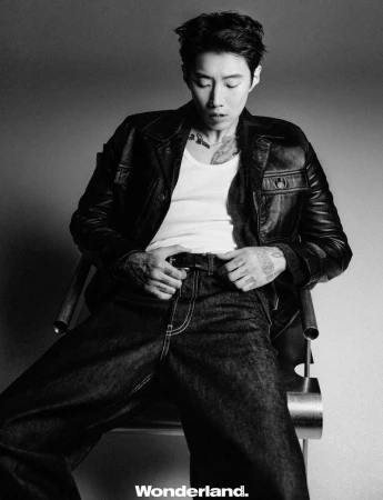 Jay Park seated in a modern chair wearing a black leather jacket, white tank top, and dark denim jeans in a monochrome studio portrait.