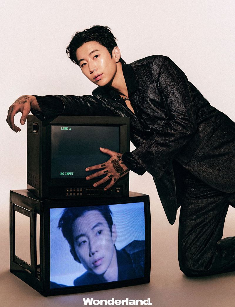 Jay Park leaning over stacked vintage CRT television monitors wearing a dark textured suit against a neutral studio backdrop.