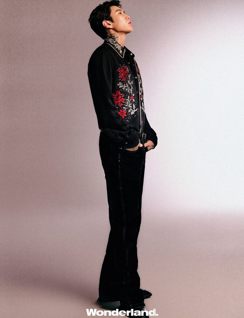 Jay Park standing in profile wearing an embroidered black shirt and black pants against a neutral studio backdrop.