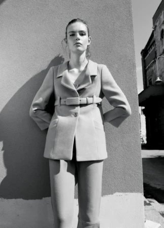 Monochrome portrait of model Anna Viksoe in a tailored belted jacket standing against an exterior wall in direct sunlight.