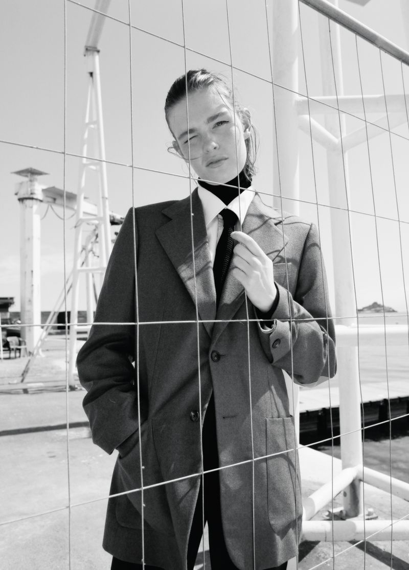 Model Anna Viksoe wearing an oversized grey blazer, white shirt, and black tie behind a wire mesh fence in black and white.