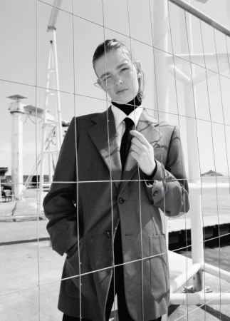 Model Anna Viksoe wearing an oversized grey blazer, white shirt, and black tie behind a wire mesh fence in black and white.