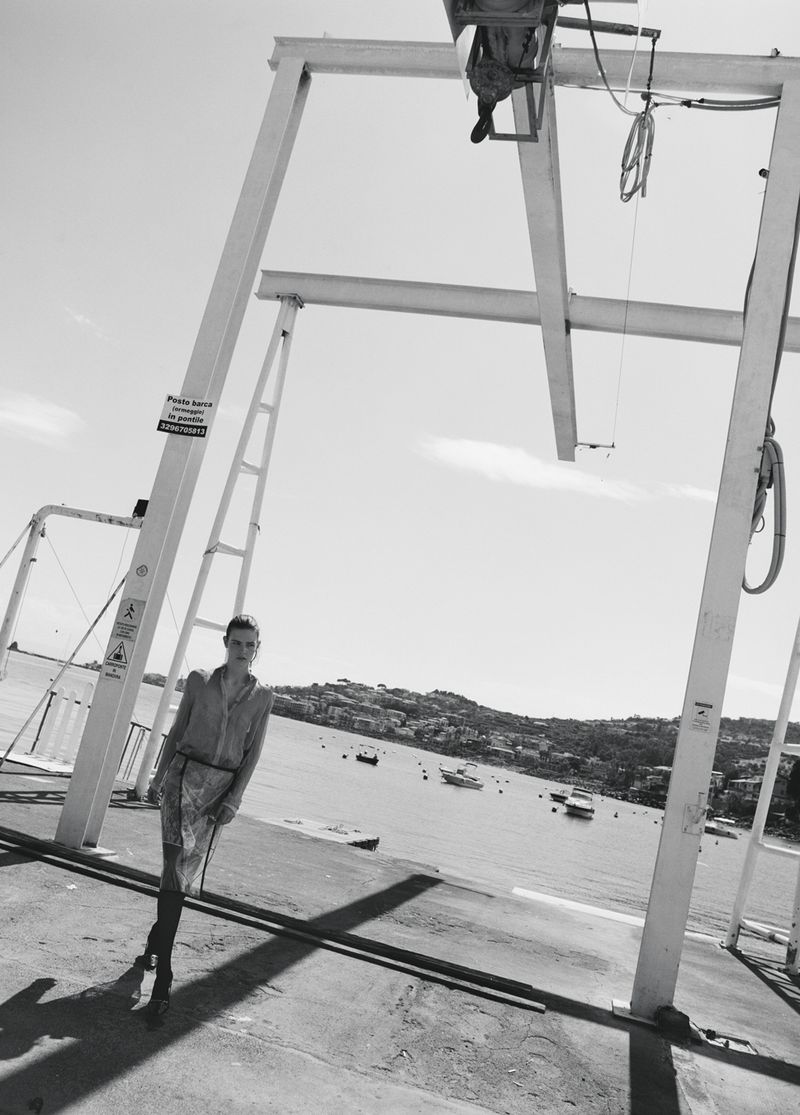 Model Anna Viksoe posing under a white industrial boat hoist on a coastal dock overlooking a bay in black and white.