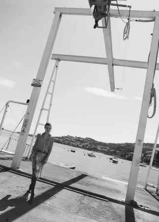 Model Anna Viksoe posing under a white industrial boat hoist on a coastal dock overlooking a bay in black and white.