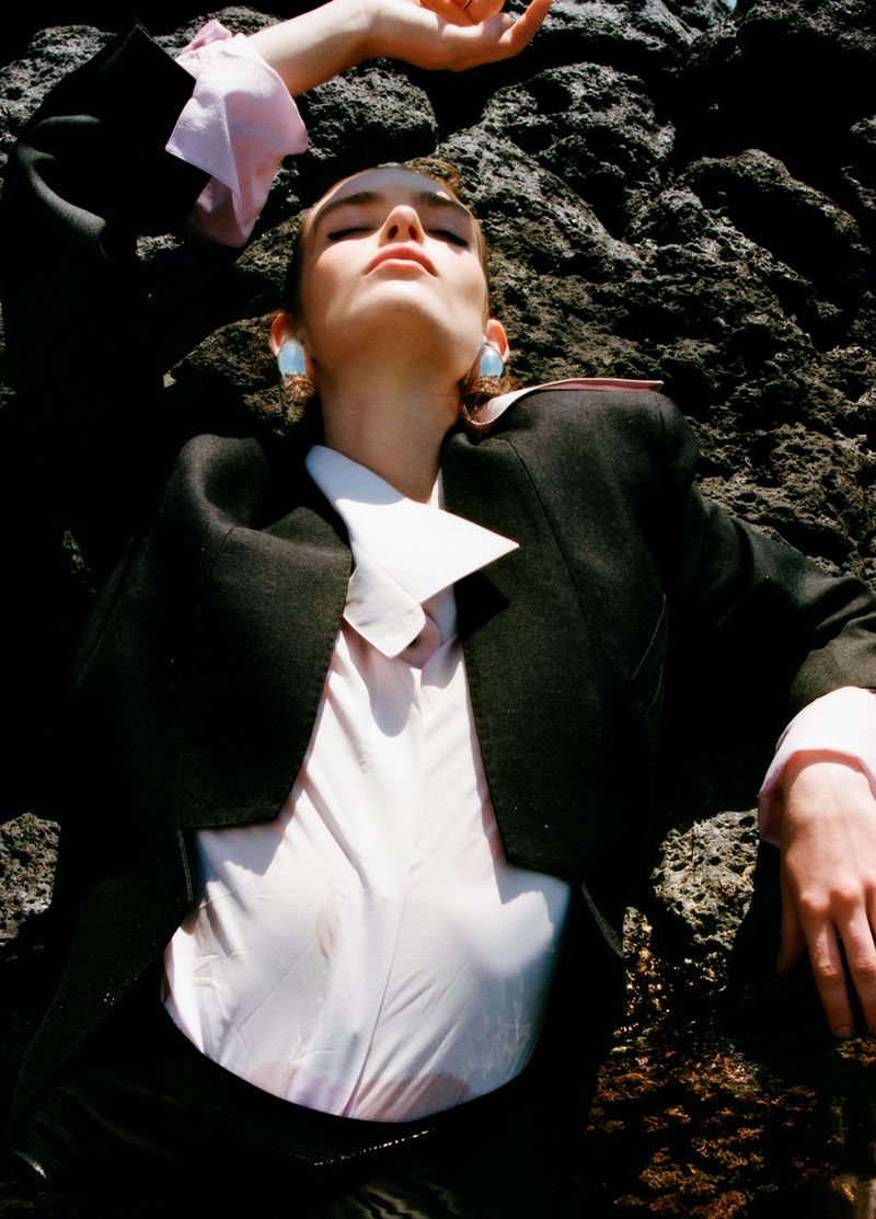 Anna Viksoe leaning back against dark volcanic rock wearing a black tailored blazer, pale pink shirt, and large statement earrings.