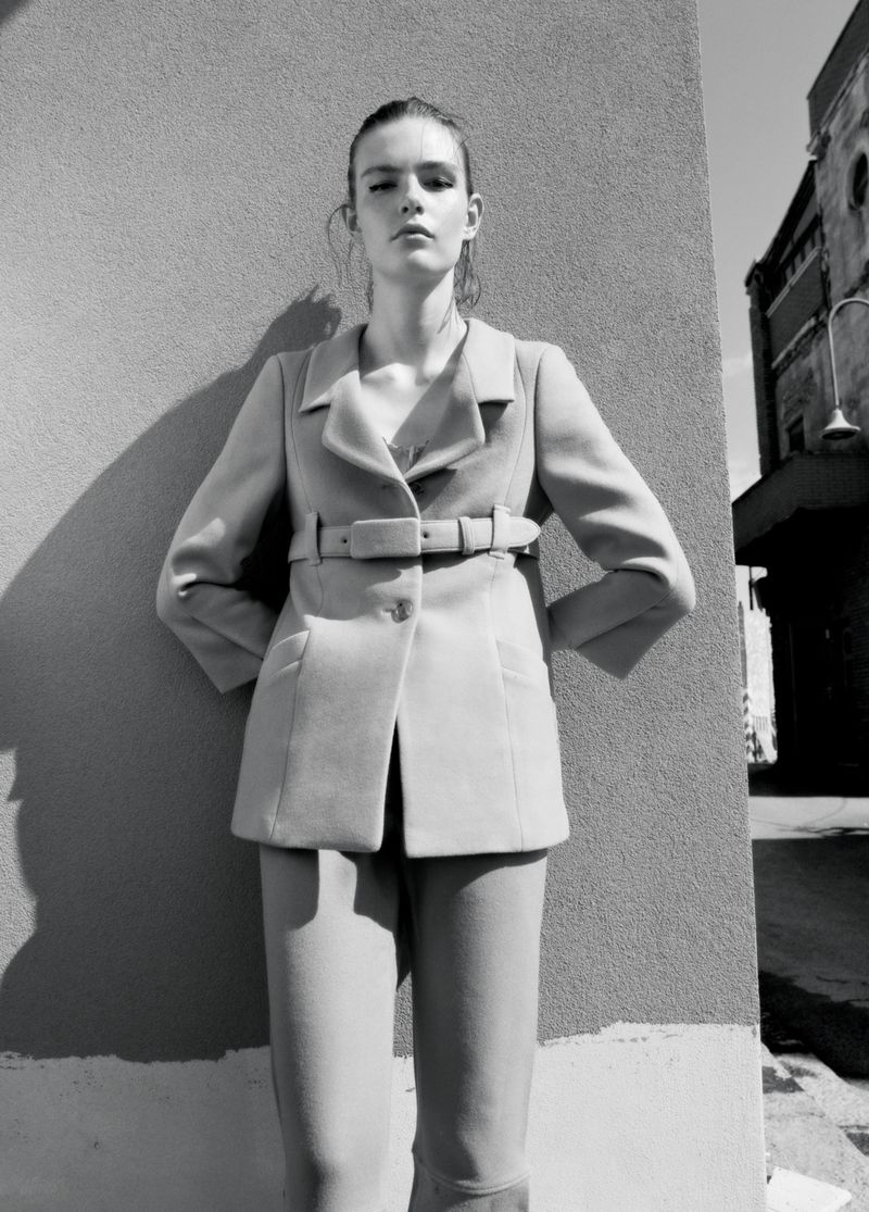 Model Anna Viksoe wearing a tailored belted jacket and trousers while posing against an outdoor wall in direct sunlight.