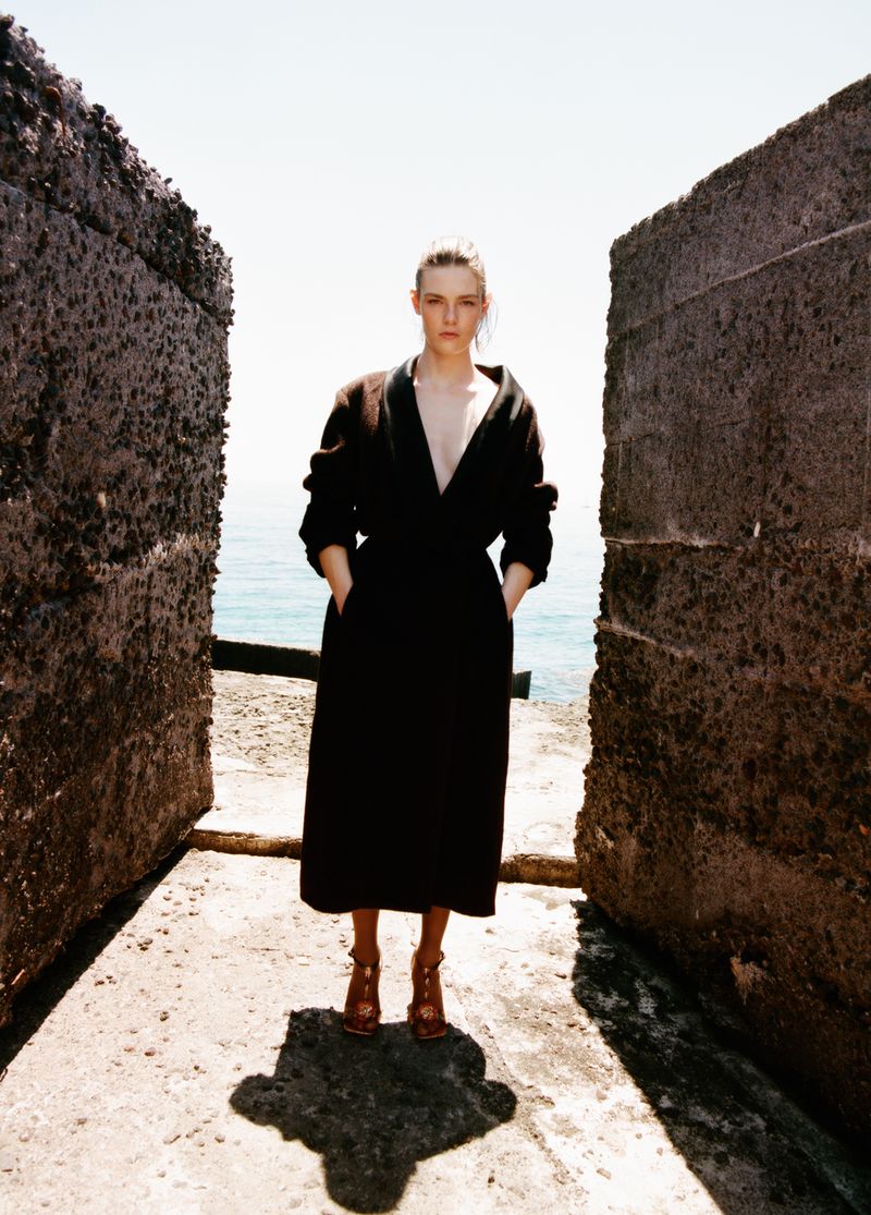 Model Anna Viksoe wearing a black V-neck coat and heels standing between concrete sea walls overlooking the ocean.