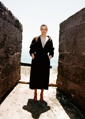 Model Anna Viksoe wearing a black V-neck coat and heels standing between concrete sea walls overlooking the ocean.