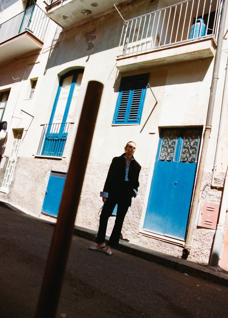 Model Anna Viksoe standing in a black suit on a sunlit street beside a building with bright blue shutters.