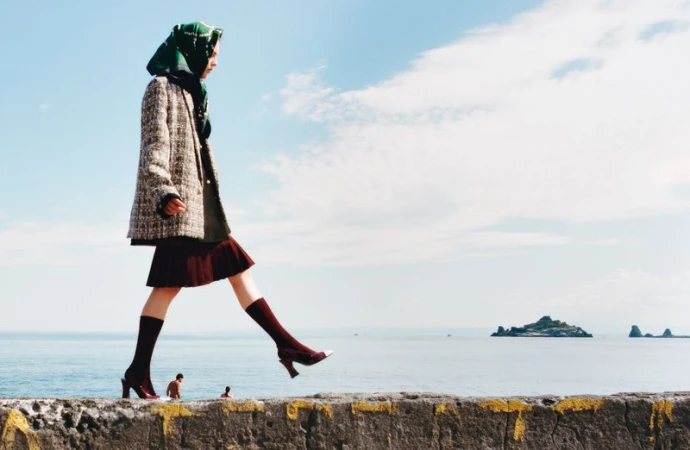 Model Anna Viksoe walks across a stone sea wall wearing a tweed blazer, burgundy pleated skirt, green headscarf, and heels by the ocean.