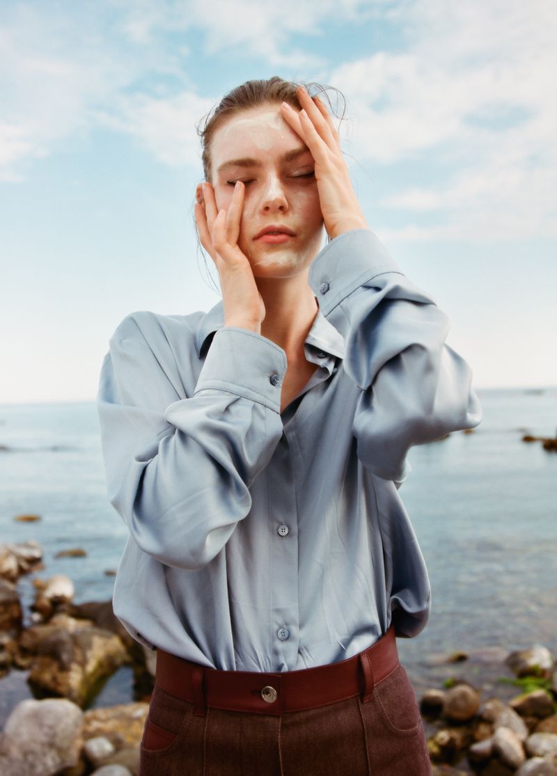 Model Anna Viksoe posing with hands on her face wearing a pale blue silk shirt and brown pants by a rocky shoreline.
