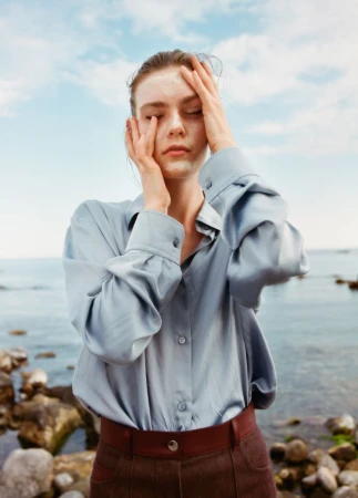 Model Anna Viksoe posing with hands on her face wearing a pale blue silk shirt and brown pants by a rocky shoreline.