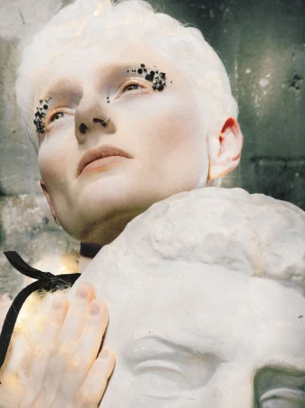 Close-up of a model with short white hair and black crystal eye makeup resting a hand on a classical plaster bust against a stone wall.