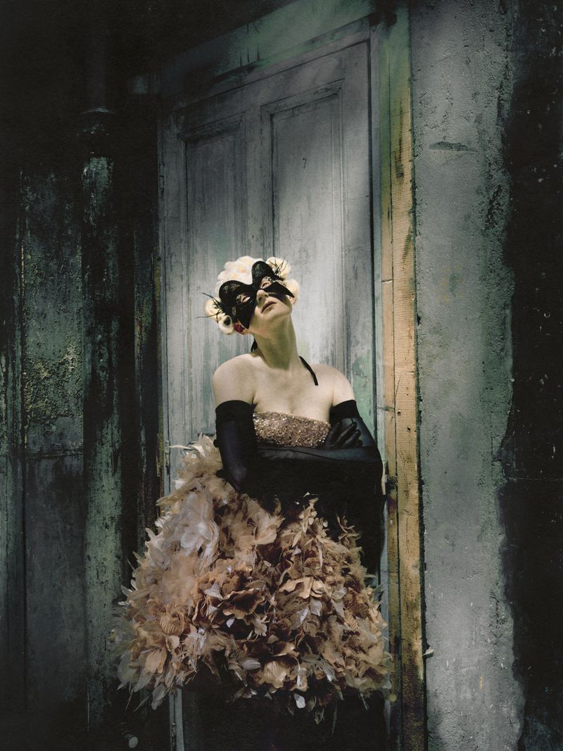 Model wearing a feathered floral skirt, black opera gloves, and an ornate mask leaning against a weathered door.