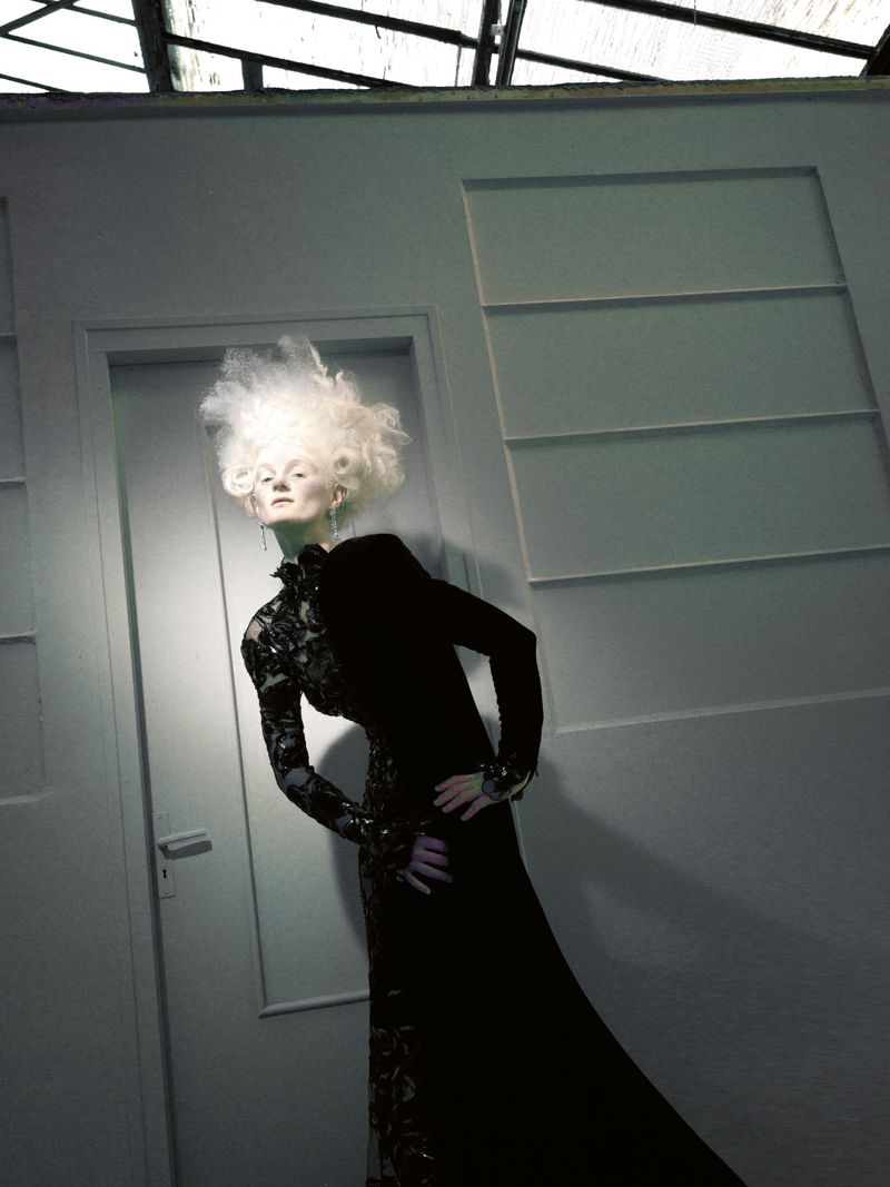 Model with dramatic white hair wearing an embellished black gown posing in an interior doorway with moody lighting.