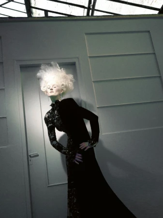 Model with dramatic white hair wearing an embellished black gown posing in an interior doorway with moody lighting.