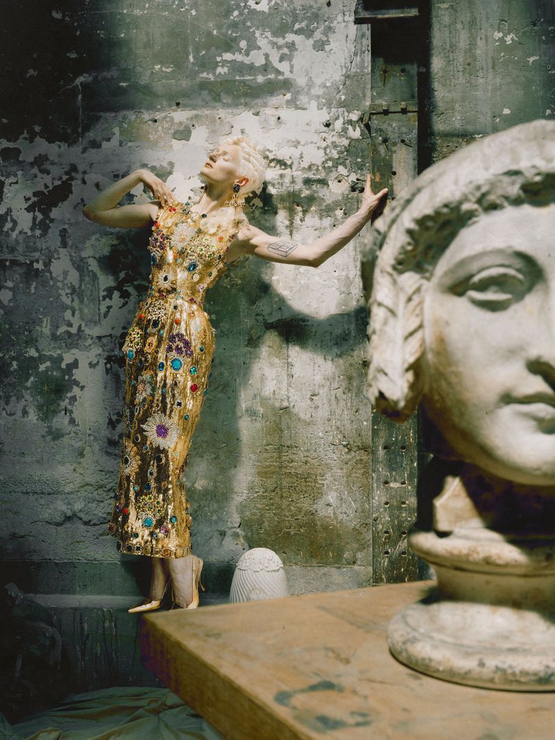 Model wearing a jeweled gold embellished dress poses against a distressed concrete wall behind a classical stone sculpture bust.