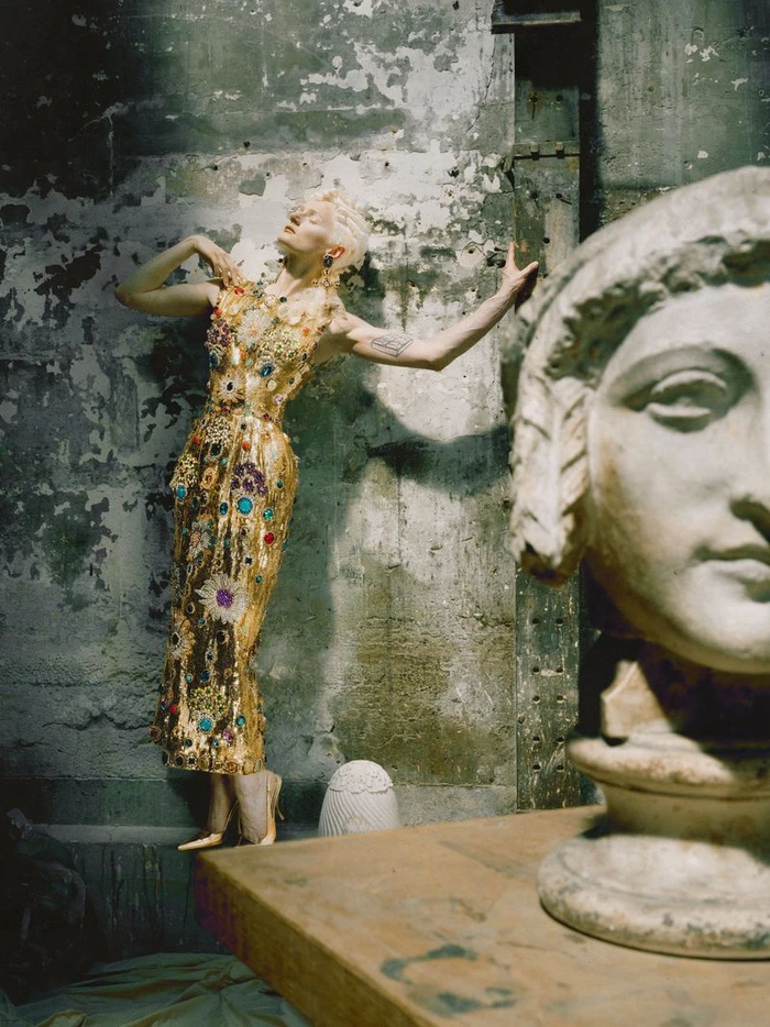 Model wearing a jeweled gold embellished dress poses against a distressed concrete wall behind a classical stone sculpture bust.