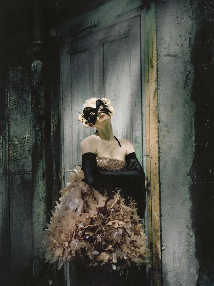 Model wearing a feathered couture dress, long black opera gloves, and a black eye mask posing against a distressed wooden door.