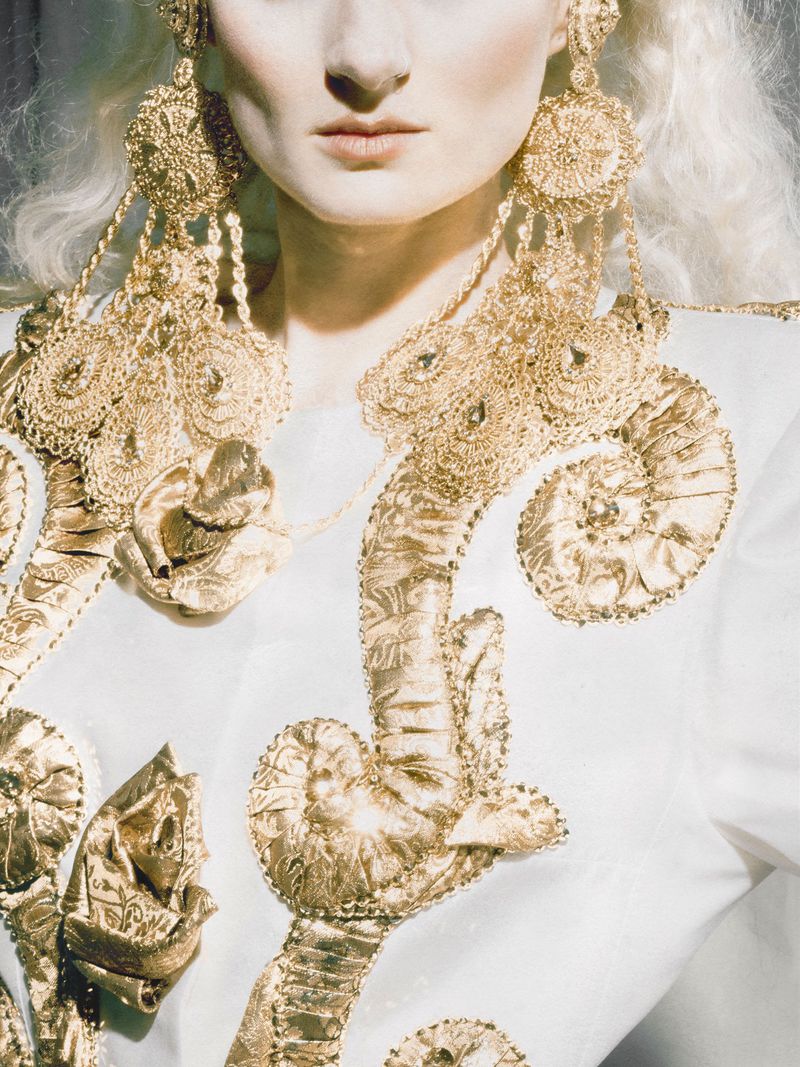 Close-up portrait of a woman wearing oversized ornate gold chandelier earrings and a white garment with gold embroidery.