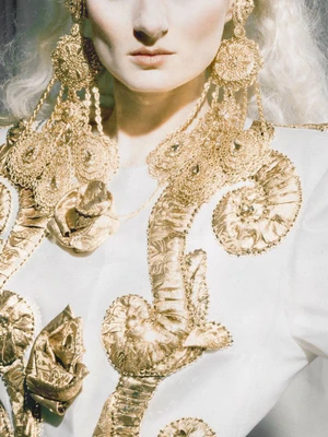 Close-up portrait of a woman wearing oversized ornate gold chandelier earrings and a white garment with gold embroidery.
