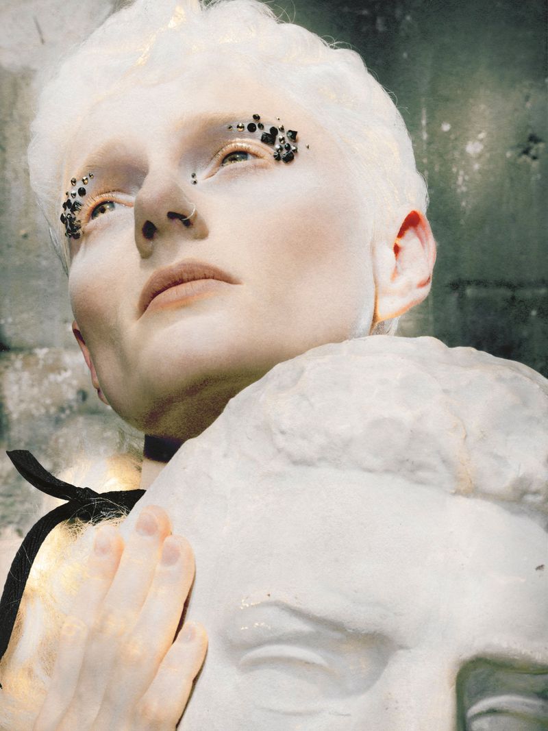 Close up of a model with white hair and rhinestone eye makeup resting their hand on a classical marble sculpture bust.